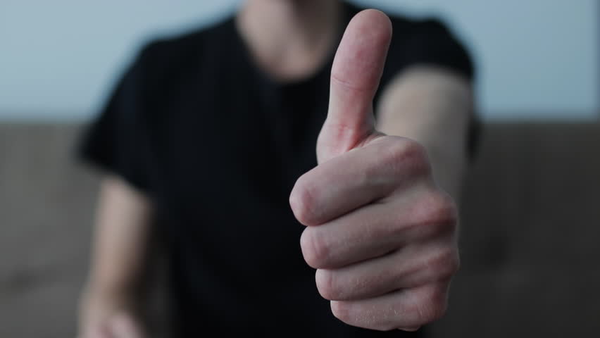 Close-up of a male hand showing a thumbs-up gesture. The young man in a black shirt expresses approval, success, and positivity