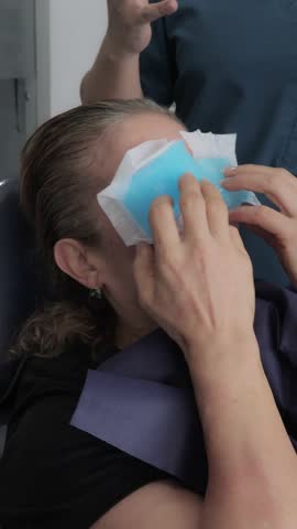 Close-up vertical shot of an elderly female patient holding a blue cold gel pack against her eyes to soothe pain and reduce swelling following an upper eyelid surgery.
