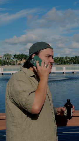Tattooed young man in casual linen shirt standing on lakeside boardwalk, talking on the phone, relaxed remote work and client communication outdoors in sunny urban park.
