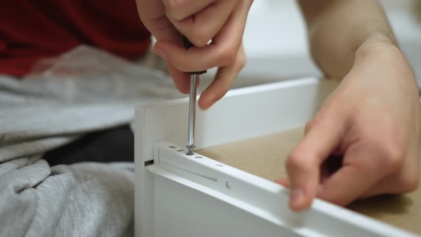 Worker tightening rail screw with screwdriver during dresser drawer assembly. Craftsman fixing slide hardware onto panel using manual screwdriver while building DIY cabinet. Carpenter securing guide