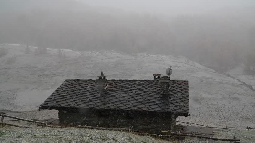 Wide shot of a rustic mountain cabin with a slate roof during a blizzard in a remote valley.