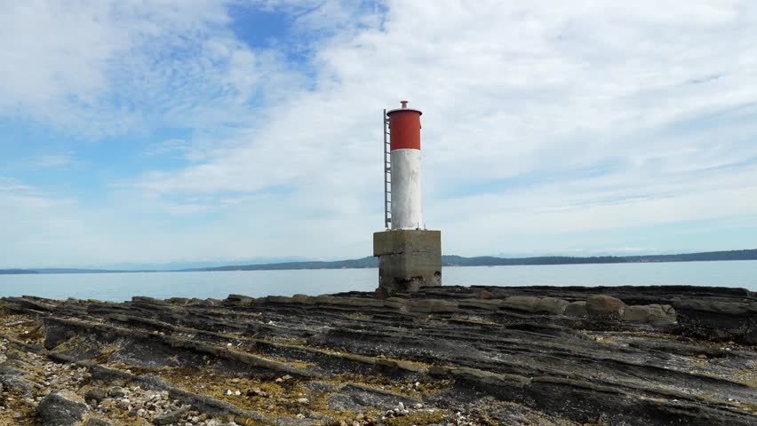 Red and white lighthouse rises from rocky shore in chemainus, british columbia, with calm ocean waters and cloudy blue sky creating a serene coastal scene