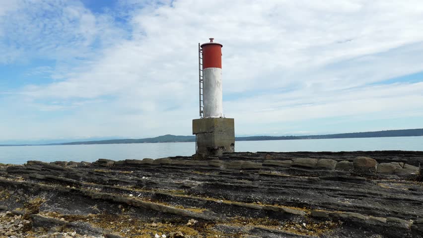 Red and white navigation marker stands on a rocky coast in chemainus, british columbia, with calm waters and a cloudy sky creating a serene maritime scene