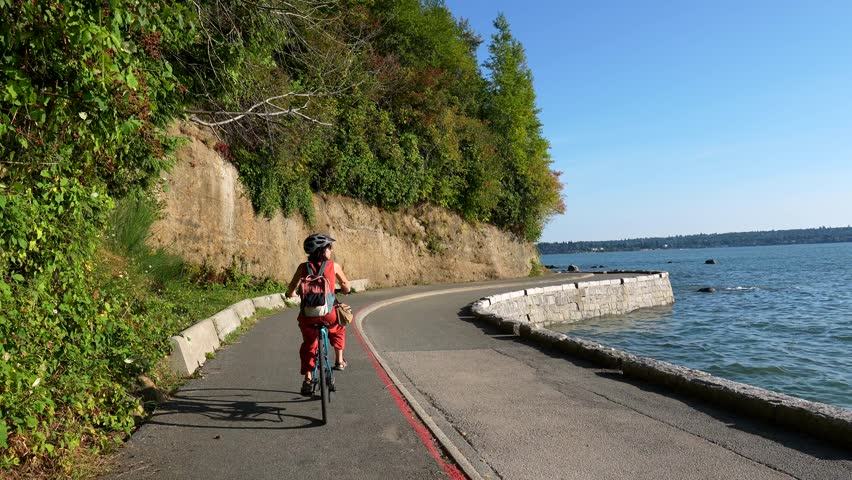 Woman cycling along scenic stanley park seawall in vancouver, enjoying ocean views, lush greenery, and coastal landscape during summer recreational bike ride