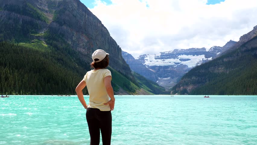 Female tourist wearing baseball cap standing at edge of turquoise lake louise, gazing at victoria glacier and surrounding mountains in banff national park, alberta, canada