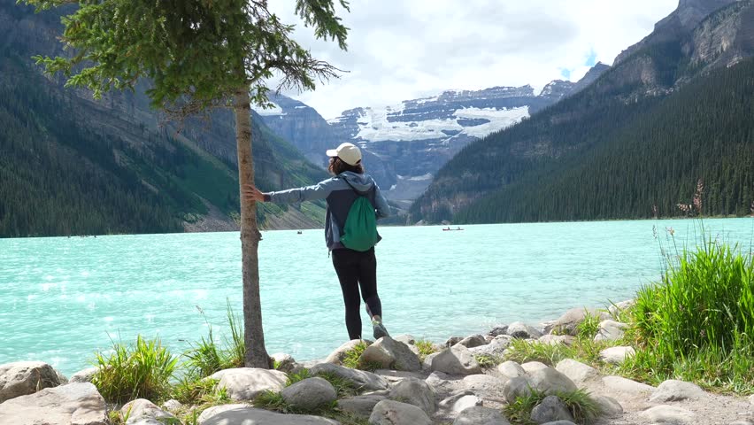 Female traveler leaning against evergreen tree, absorbing panoramic mountain landscape with turquoise lake louise and victoria glacier in background of banff national park