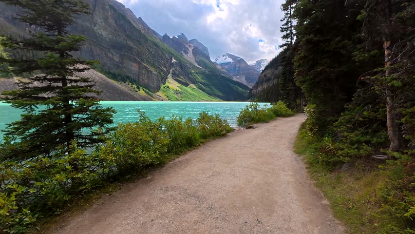 Turquoise water of lake louise laps the shoreline trail, offering breathtaking views of the surrounding canadian rockies, with snow capped peaks and lush greenery under a cloudy sky