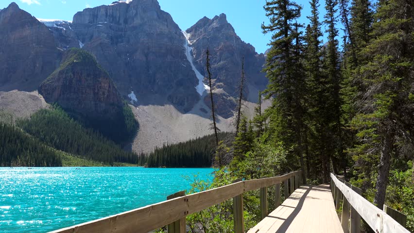 Turquoise water reflects the surrounding mountains and forests as someone walks along a wooden path by moraine lake in banff national park, offering a serene view of nature