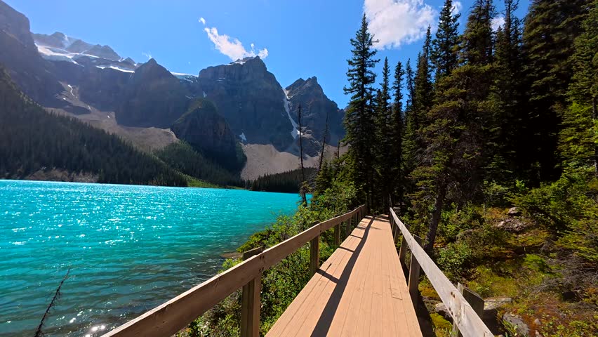 Wooden boardwalk traversing turquoise moraine lake, reflecting majestic mountains and verdant forest under bright summer sky in banff national park, alberta, canada