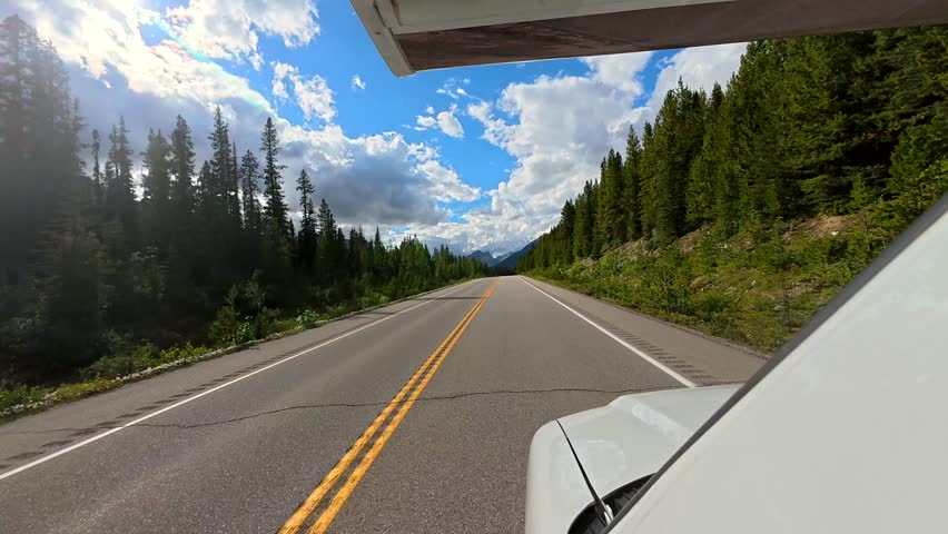 White motorhome driving on a scenic highway through the canadian rockies, offering breathtaking views of the mountains and forest landscape under a summer sky