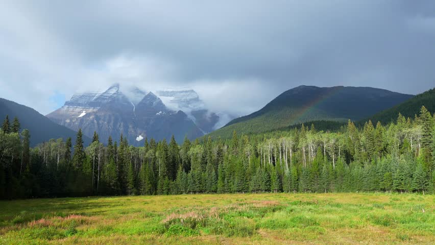 Clouds envelop mount robson, the highest peak in the canadian rockies, while a vibrant rainbow arches over a lush green meadow, creating a breathtaking panorama of nature