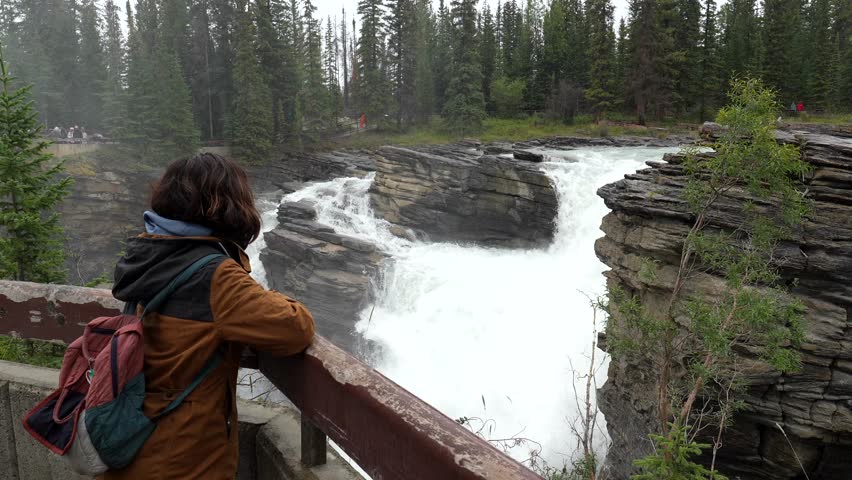 Young female tourist wearing a brown jacket and backpack leaning on a wooden railing, contemplating athabasca falls in jasper national park, canada