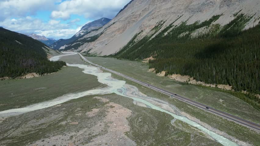 Several cars are driving on the icefields parkway next to a glacial river with a mountain and forest in the background near the columbia icefield in jasper national park, alberta, canada