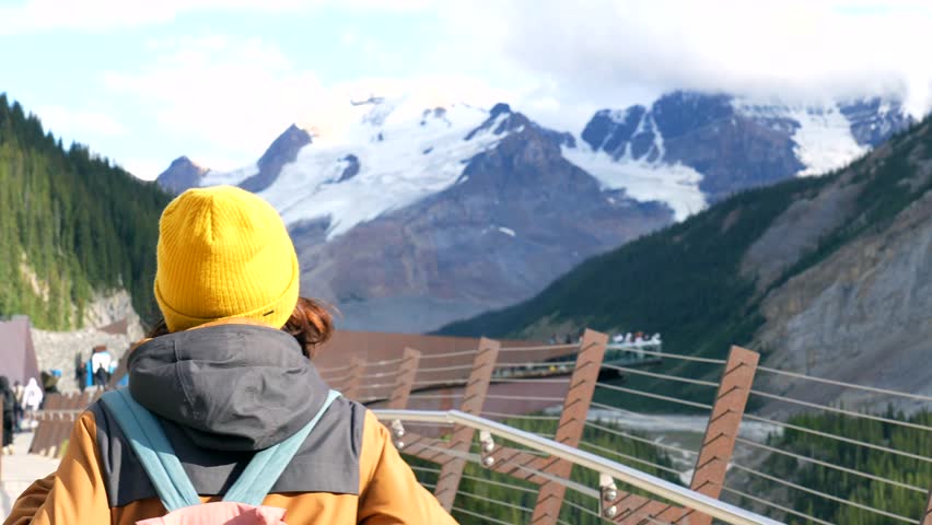 Hiker wearing yellow hat and backpack walking on skywalk at columbia icefield, viewing athabasca glacier and surrounding canadian rockies mountains in jasper national park, alberta