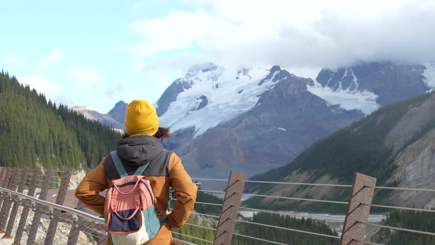 Female traveler wearing yellow beanie and backpack standing on glacier skywalk, admiring panoramic views of athabasca glacier in jasper national park, alberta, canada