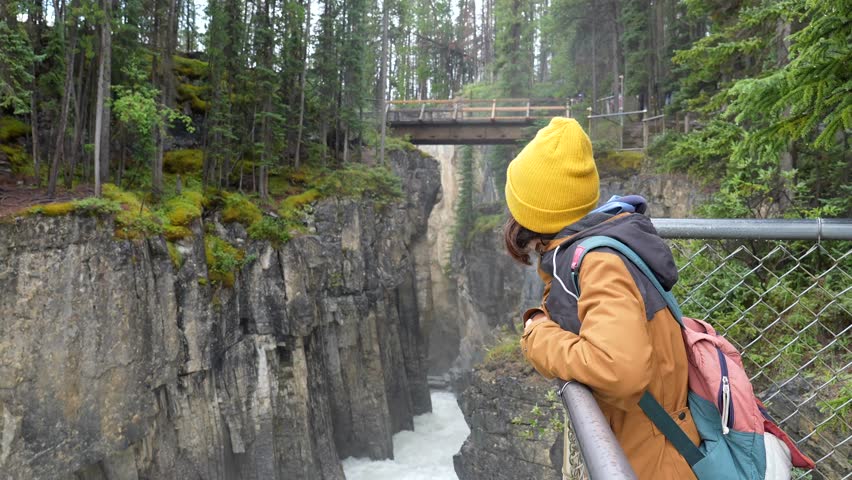 Female tourist wearing yellow beanie and brown jacket leaning on metal railing, admiring sunwapta falls and wooden bridge in jasper national park, canadian rockies