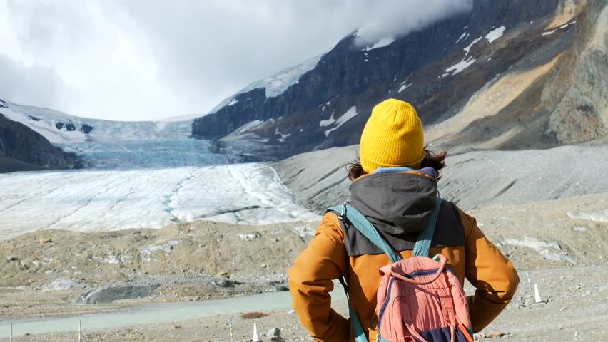 Young woman tourist with backpack and yellow hat looking at athabasca glacier in columbia icefield in jasper national park, alberta, canada