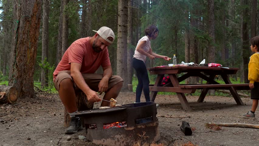 Tourists are enjoying cooking on a fire pit in banff national park, canada, surrounded by a lush forest, with father grilling and children arranging food on a picnic table