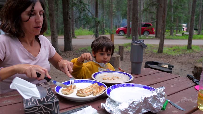 Happy family enjoys a meal together at a picnic table in banff national park, with a mother carefully cutting grilled chicken for her young son
