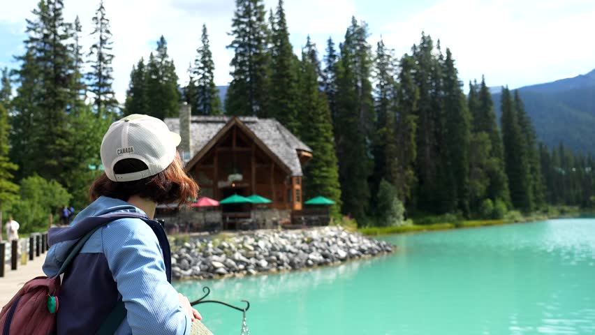 Female tourist wearing a cap and backpack admires the turquoise waters of emerald lake and its wooden lodge in banff national park, yoho national park, british columbia, canada