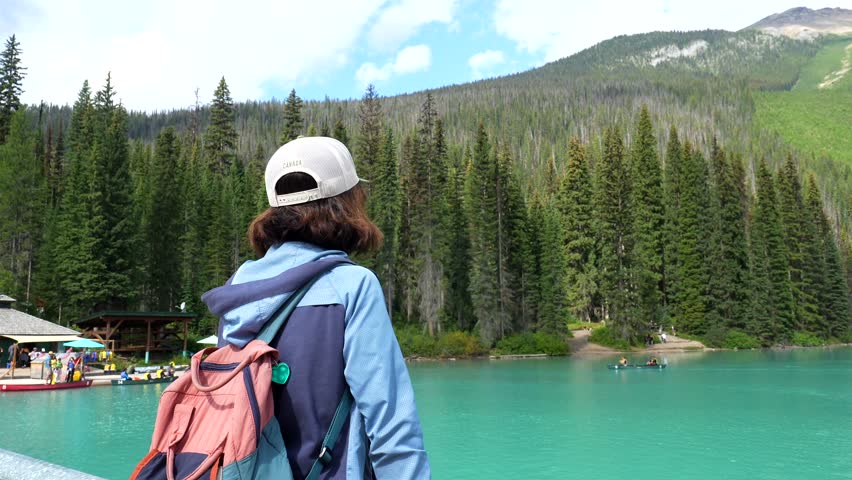 Female tourist wearing a cap and backpack admires the stunning turquoise waters of emerald lake, surrounded by a dense coniferous forest and mountain range in banff national park, canada