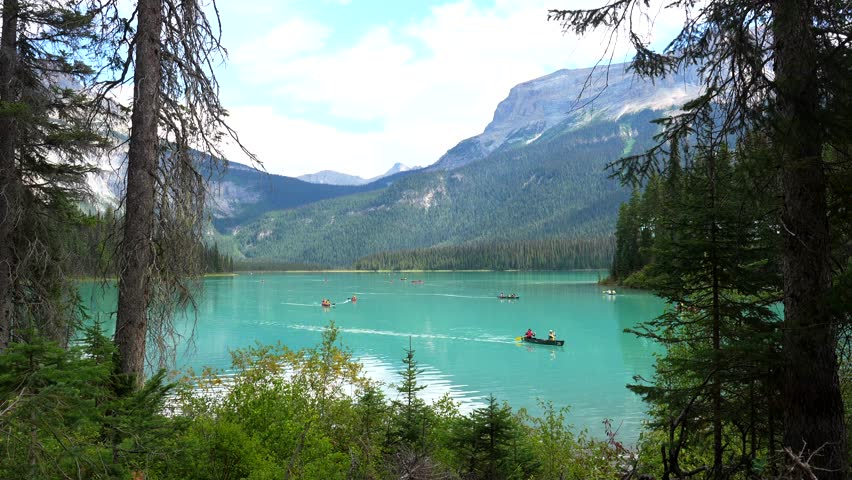 Emerald lake, nestled in the canadian rockies, hosts tourists canoeing across its turquoise waters, surrounded by lush forests and majestic mountains under a summer sky