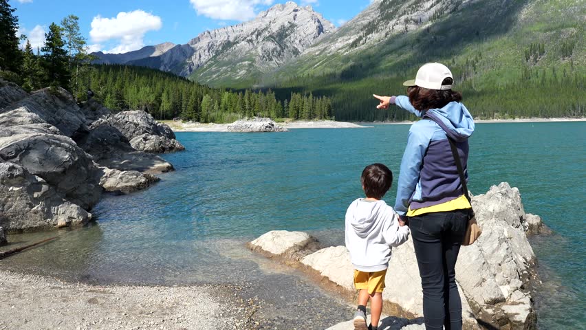 Mother and son standing on rocky shore of lake minnewanka, pointing towards distant mountain ranges in banff national park, sharing moment of discovery amid pristine canadian wilderness