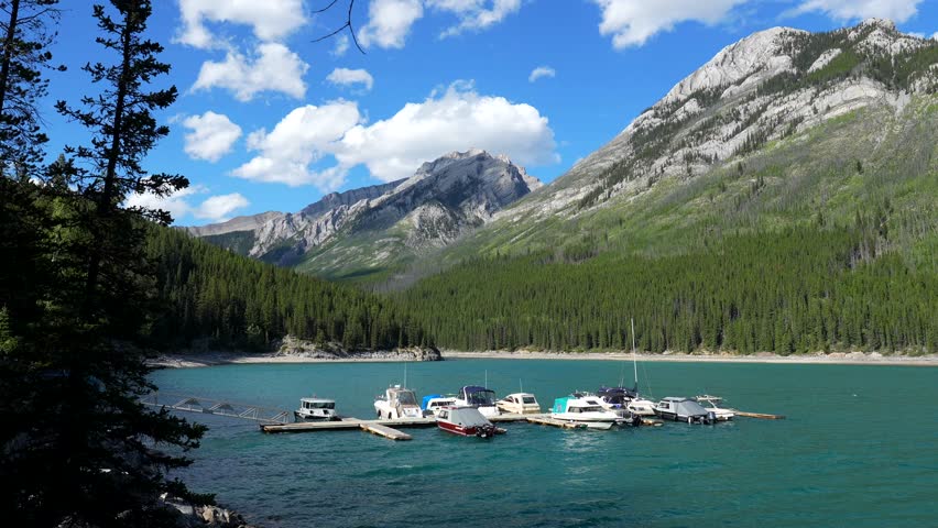 Azure water reflects the sky and clouds, mirroring the majestic mountains and lush green forests surrounding lake minnewanka in banff national park, where several boats rest peacefully at a dock