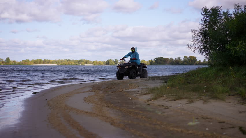 Slow motion. Rider on ATV tracks along lakeshore sand and mud, yellow jacket and helmet, tight cornering leaving curved tire marks, water splashes and wet shoreline, cloudy.