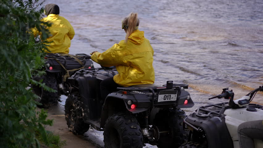 Slow motion. Yellow jacket riders preparing for ride, two riders in bright rain coats mounting quads at muddy shore, checking helmets and revving engines, energetic prelude