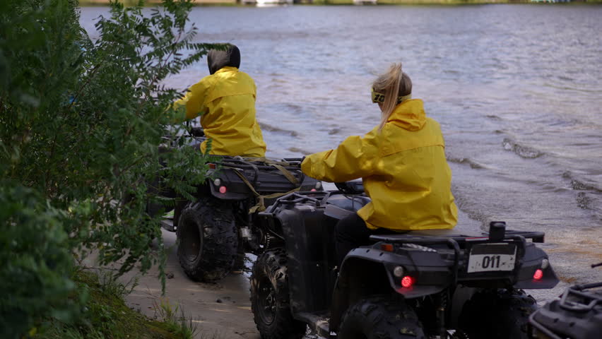 Slow motion. Riders wave on shore before departure, two companions in yellow jackets exchanging goodbye wave while turning ATVs away from lake, gentle splash and serene