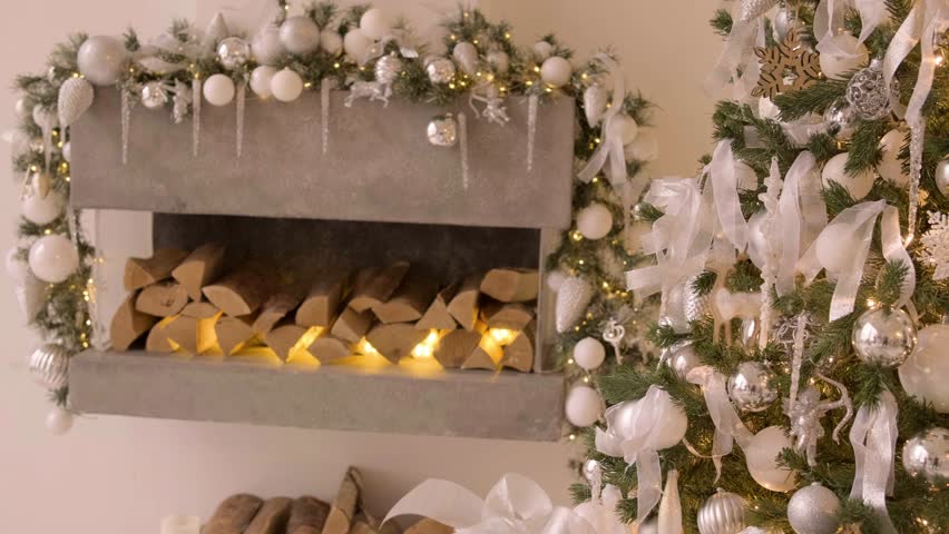 Closeup of white Christmas tree decorated with festive lights and ornaments, wrapped white gifts nearby and fireplace in background. White color of year