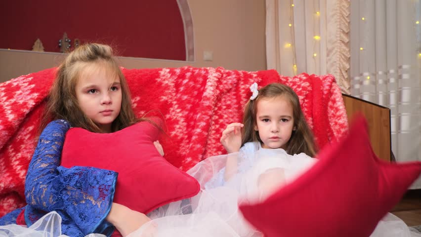 Two sisters in festive dresses sitting in the bedroom decorated with a Christmas tree and a garland
