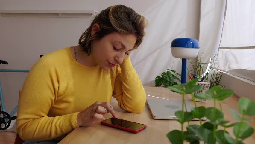 Uninterested young woman in a yellow sweater endlessly scrolling on her mobile phone screen, procrastinating while sitting at her home office desk next to a closed laptop and a plant