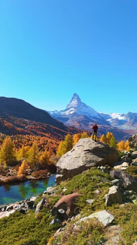 Hiker Man on Top of Boulder Looking at Matterhorn Mountain. Lake Grindjisee and Yellow Larches in Autumn. Fall Colors. Swiss Alps. Aerial View. Zermatt, Valais, Switzerland. Vertical Video