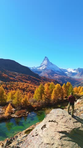 Hiker Man on Top of Boulder Looking at Matterhorn Mountain. Lake Grindjisee and Yellow Larches in Autumn. Fall Colors. Swiss Alps. Aerial View. Zermatt, Valais, Switzerland. Vertical Video