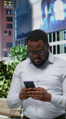 Vertical Video Adult man looks for directions on phone gps and walks across city pavement, modern sense of style and city life. Black person strolling and checking his smartphone apps, urbanism