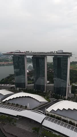 Vertical aerial view of Marina Bay Sands in Singapore, showing the three iconic towers with rooftop sky park, modern architecture, waterfront, greenery, and ships anchored in the bay.