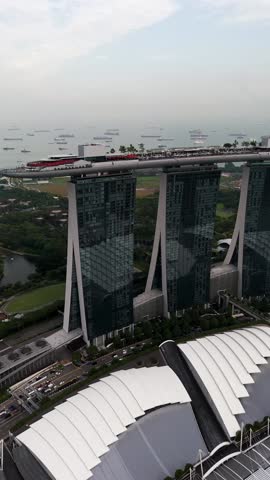 Vertical aerial view of Marina Bay Sands in Singapore, featuring the three towers and rooftop SkyPark, surrounding greenery, waterfront structures, and cargo ships anchored in the bay.