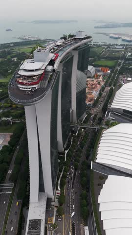 Vertical aerial view of Marina Bay Sands in Singapore, highlighting the SkyPark rooftop, luxury hotel towers, surrounding roads, green areas, construction zones, and ships in the bay.