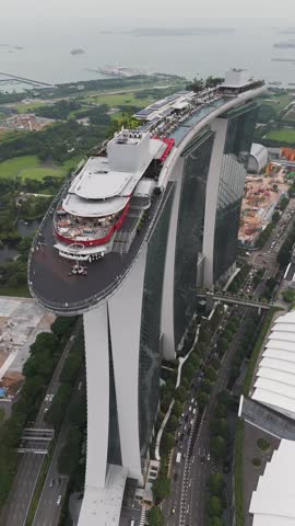 Vertical aerial view of Marina Bay Sands in Singapore, focusing on the SkyPark rooftop, infinity pool area, luxury hotel towers, surrounding roads, greenery, and ships anchored in the bay.