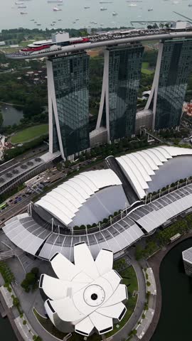 Vertical aerial view of Marina Bay Sands in Singapore, highlighting the three hotel towers, SkyPark rooftop with infinity pool, surrounding roads, greenery, waterfront, and anchored ships.