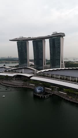 Vertical aerial view of Marina Bay Sands in Singapore, highlighting the three hotel towers, SkyPark rooftop with infinity pool, surrounding roads, greenery, waterfront, and anchored ships.