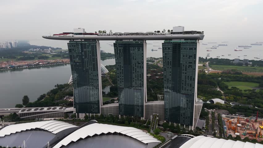 Aerial view of Marina Bay Sands in Singapore, featuring the iconic three towers connected by the SkyPark rooftop, surrounded by greenery, waterfront, urban roads, and anchored ships.