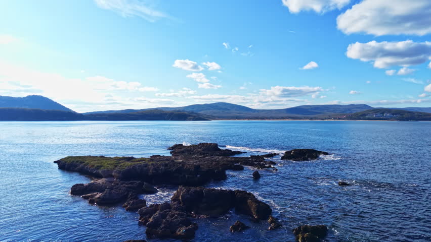 Glimmering waves lap against rugged rocks as the sun shines brightly. Rolling hills and distant mountains frame the picturesque coastal view, creating a sense of peace.
