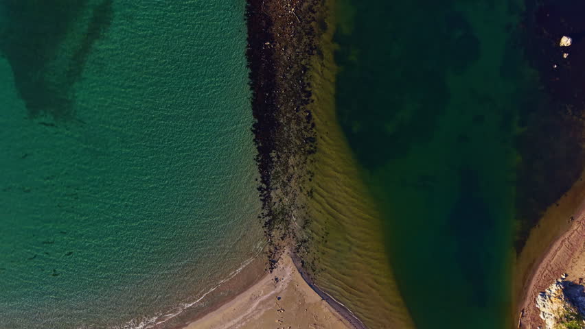 Waves gently lap against the sandy beach as vibrant turquoise waters meet deeper greens. Shadows dance along the rocky edge under the bright afternoon sun, creating stunning contrast.