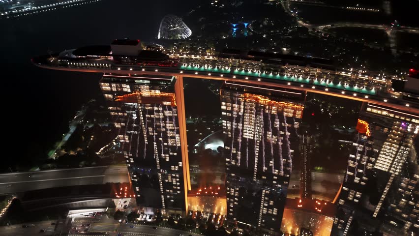 “Night aerial view of Marina Bay Sands in Singapore, illuminated three towers connected by the SkyPark rooftop, city lights, waterfront reflections, and ships in the bay.

