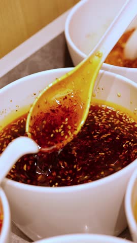 Hand stirs vibrant chili oil with sesame seeds in white bowls under warm indoor lighting