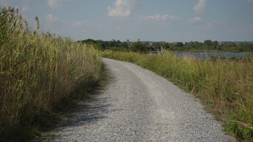 Curving Gravel Path Through Marsh Grasses Beside Quiet Lagoon Under Blue Sky, Inviting Walking Route And Coastal Atmosphere