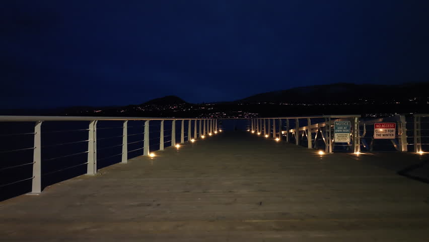 A night time view of an empty lit up boardwalk disappearing into view into a lake, with mountains in the background. 
